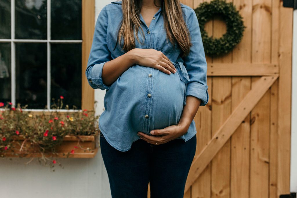 A pregnant woman wearing a blue denim shirt, standing outdoors in front of a a window and a barn-style door. A windowbox of flowers is out of focus in the background. Her hands are resting above and below her belly. Image credit: Camylla Battani via Unsplash.