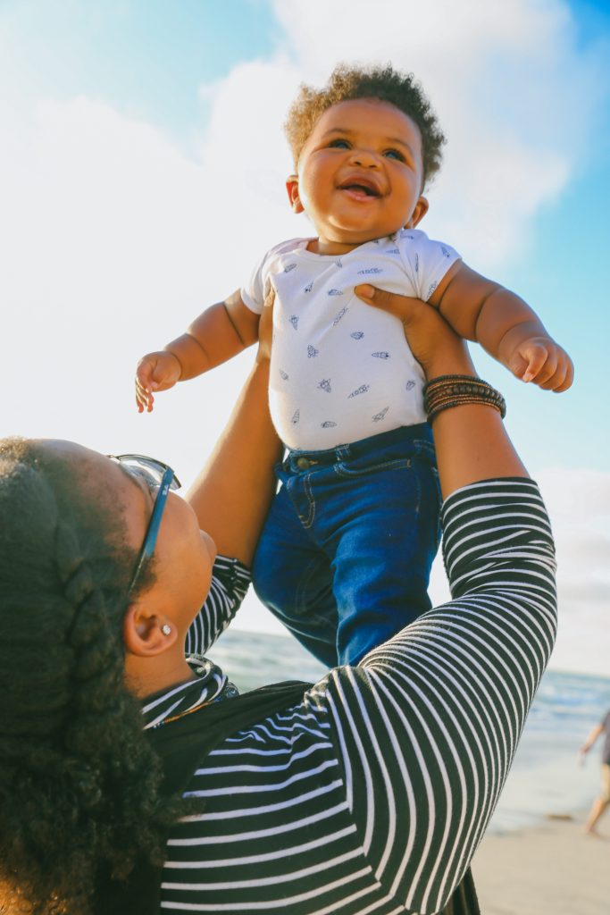 A Black mother, back partially turned to the camera, is at the beach on a sunny day. She is holding a happy, laughing infant up above her head. Image credit: Lawrence Crayton via Unsplash.
