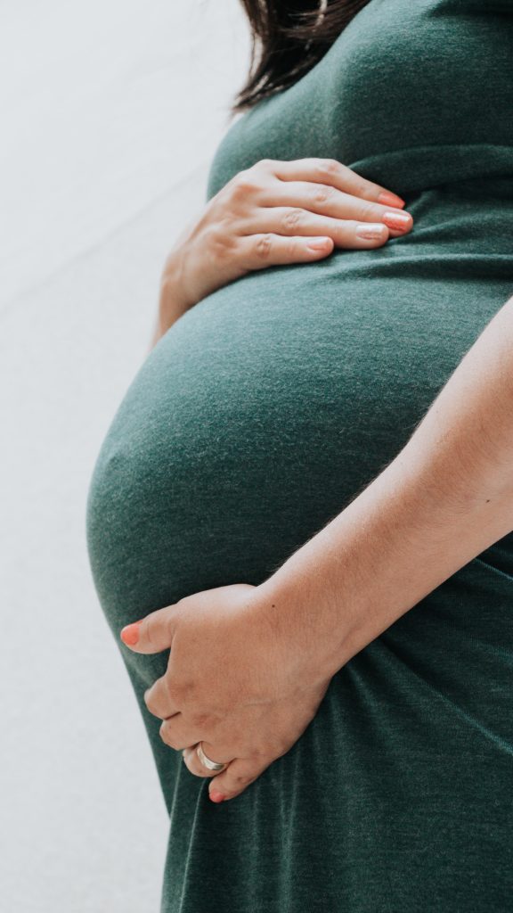 A cropped photograph of a pregnant woman, standing in semi-profile, with hands resting above and below her belly. Image credit: Vanessa via Unsplash.