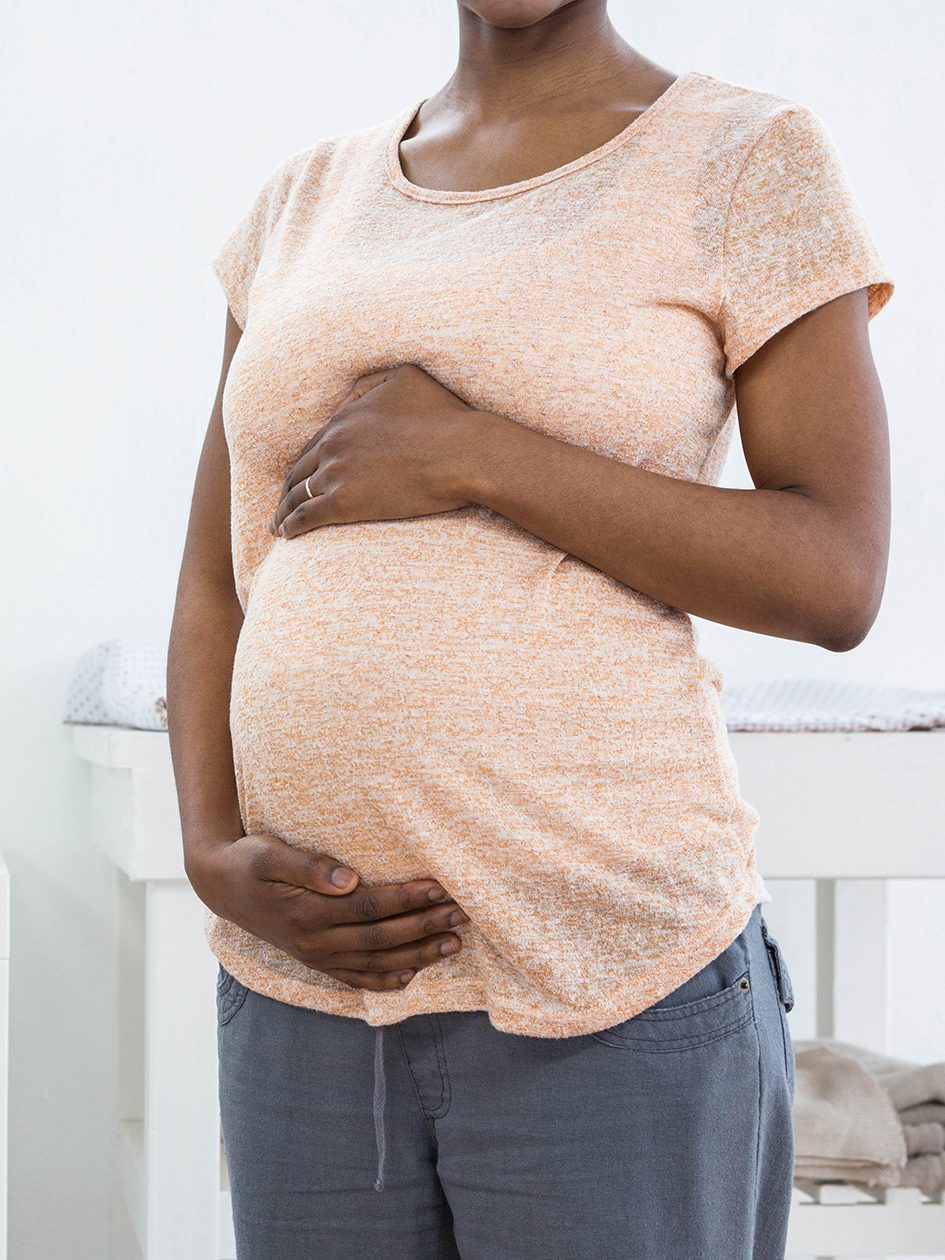 A pregnant Black woman standing infront of cradle, with hands placed above and below her stomach.