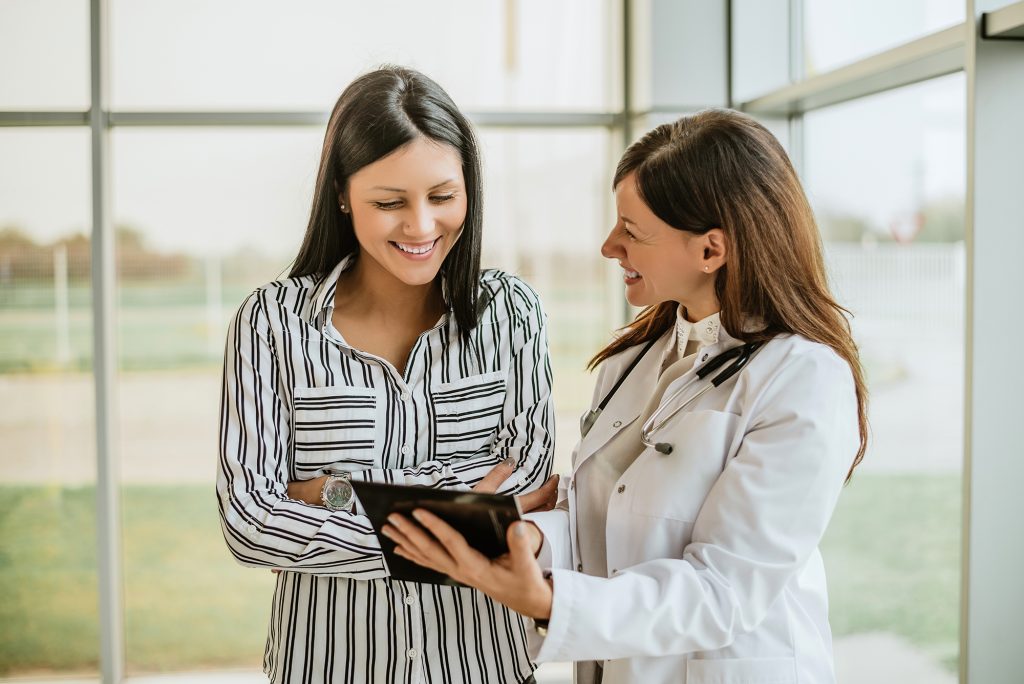 A female doctor and her female patient, both smiling, look at information the doctor is showing the patient on a tablet computer.