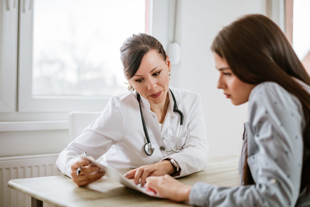 A female doctor in white coat sits at at table with her female patient. The doctor is pointing out some information contained in a chart that the two of them are looking at.