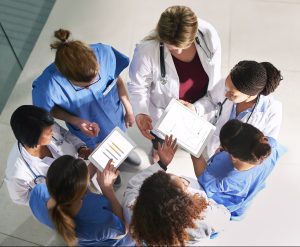 Photograph taken from above showing a tight circle of female health care providers, some in white coats and some in blue surgical scrubs, looking at and discussing data displayed on tablet computers.