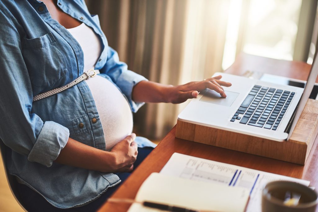 Photograph of a pregnant woman, with one hand cradling her belly, the other using a laptop computer in a home office.