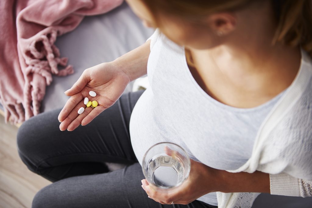 A high-angle photo, looking down at a pregnant woman dressed in jeans, t-shirt and sweater. She is holding and looking at a handful of yellow and whie pills. She holds a glass of water in the other hand.