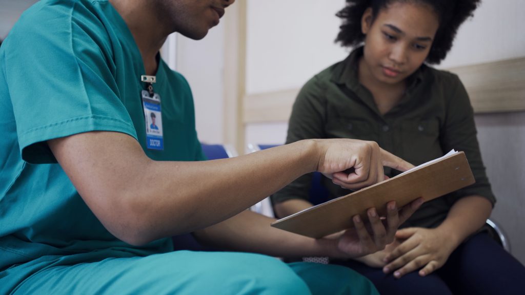 Medical assistant in scrubs with nametag is sitting with a female patient while they both look at a paper on a clipboard and the medical assistant points to the paper